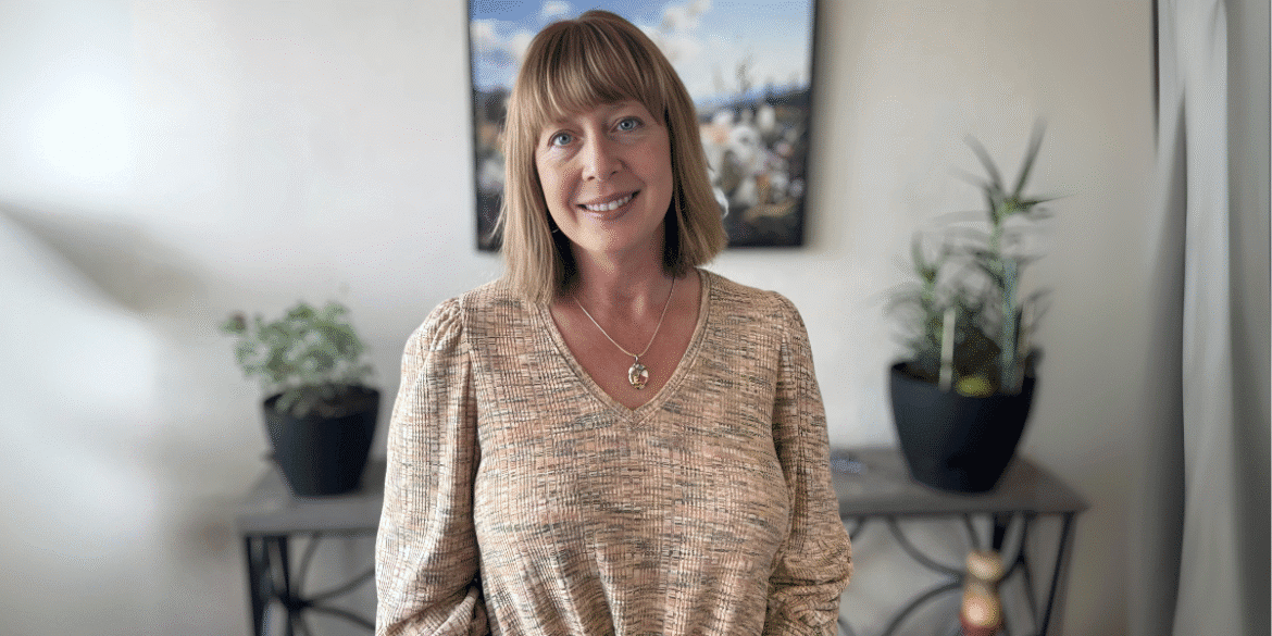 "A woman standing in front of a plant-filled table, smiling warmly. She wears a textured beige top and a pendant necklace. The background has a scenic photograph of nature."