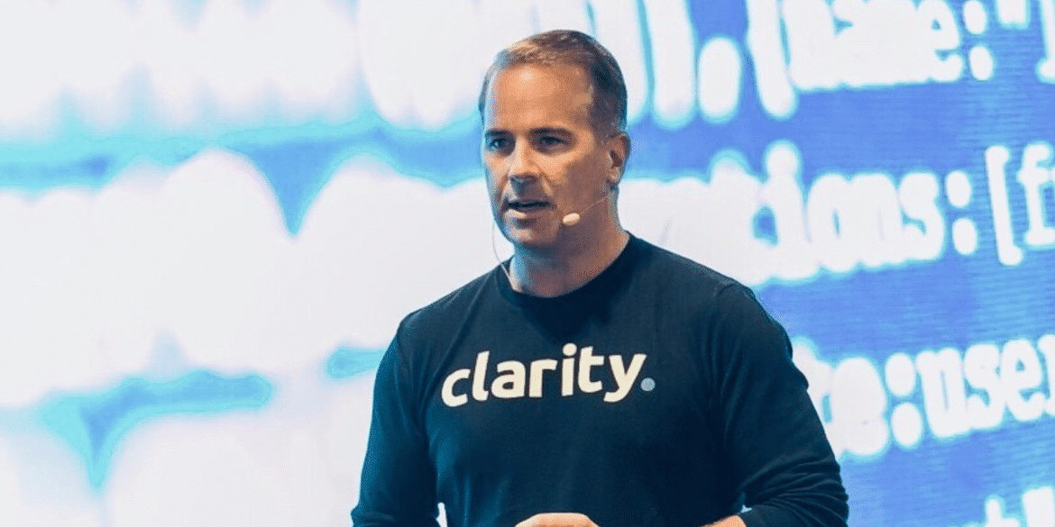 Man speaking at a conference wearing a 'clarity' branded shirt, with a digital background.