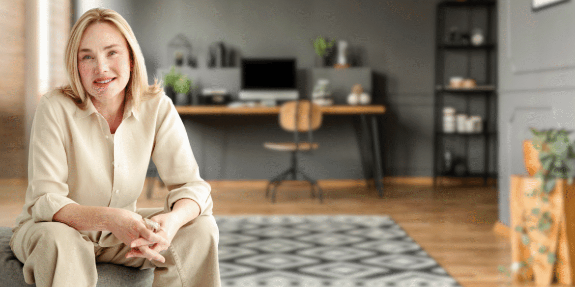 "Smiling woman in a beige blouse and pants, sitting comfortably on a chair in a modern, stylish home office with a wooden desk, computer, and plants in the background."