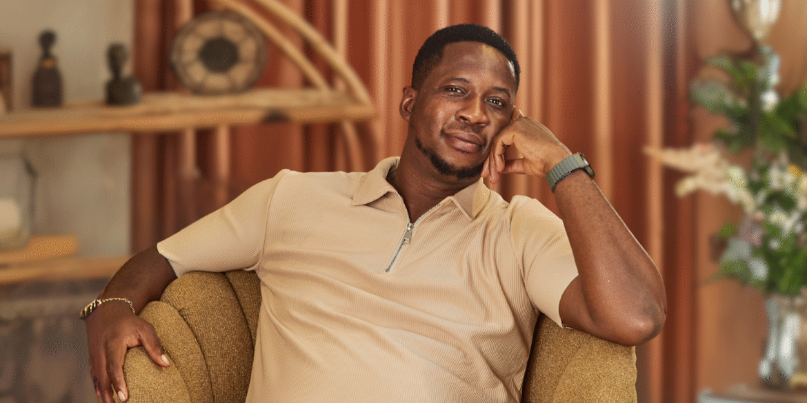"A confident man in a beige polo shirt smiles while resting his head on his hand, seated comfortably on a stylish armchair in a modern, cozy interior with soft lighting and decorative plants."