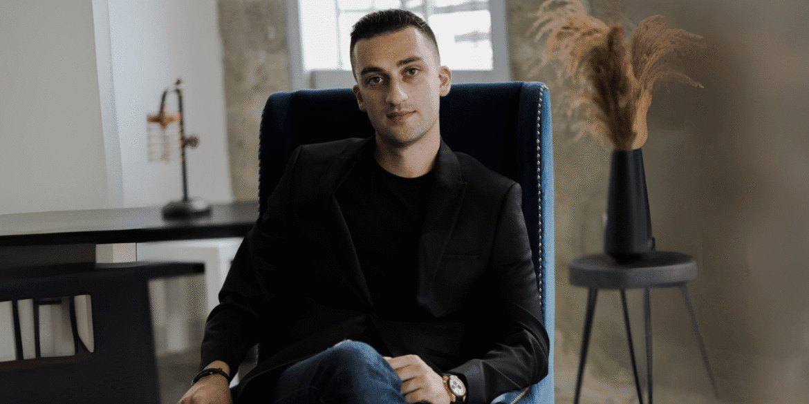 A man in a black blazer and jeans sitting confidently in a blue chair with a minimalist office background.