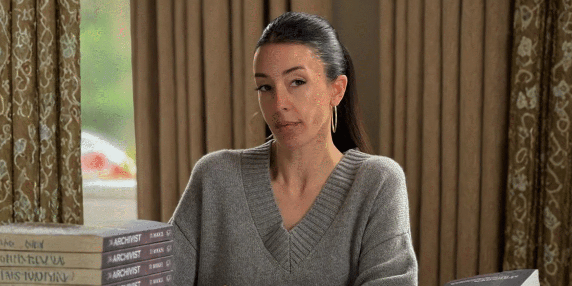 A woman in a gray sweater with hoop earrings, sitting at a table with copies of 'The Archivist' by Ti Mikkel, looking thoughtfully at the camera.