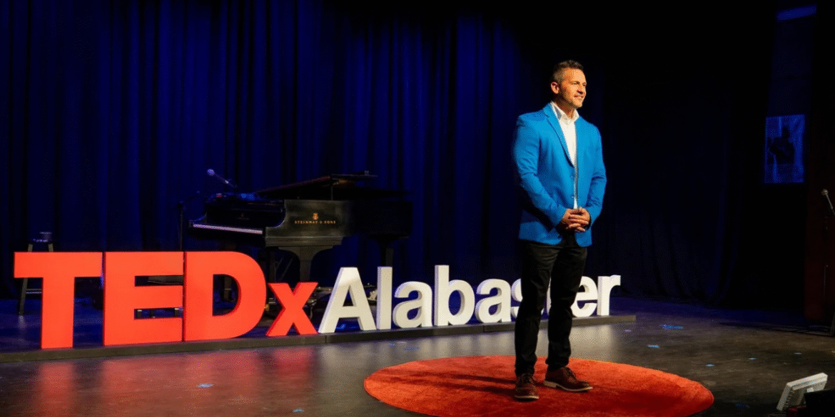 A man in a blue blazer stands on stage at TEDxAlabaster, with a piano and TEDx sign in the background.