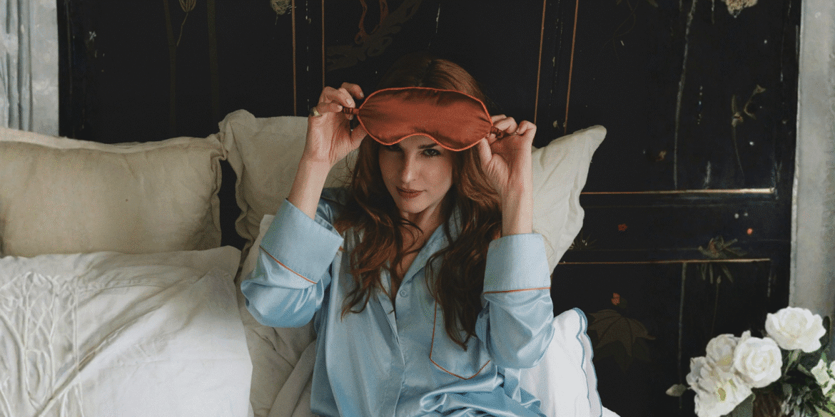 A woman in light blue silk pajamas holds a sleep mask, sitting on a bed surrounded by soft pillows and flowers.