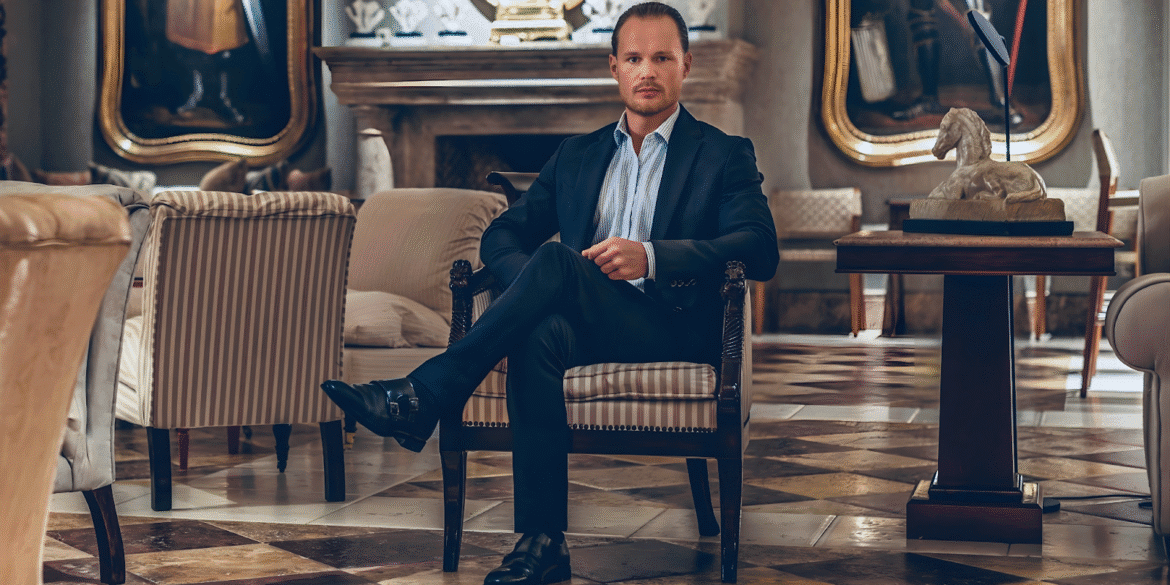 A confident man in a tailored dark suit sits cross-legged in an elegant, classical lounge with marble floors and antique décor.