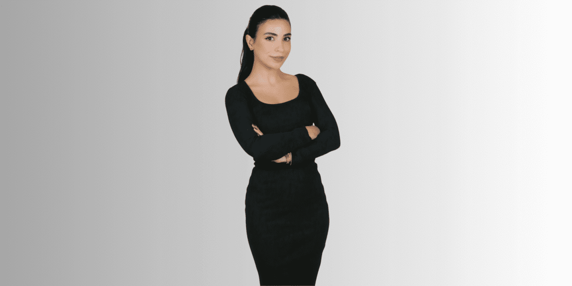 Woman in black dress standing confidently with arms crossed against a neutral background.