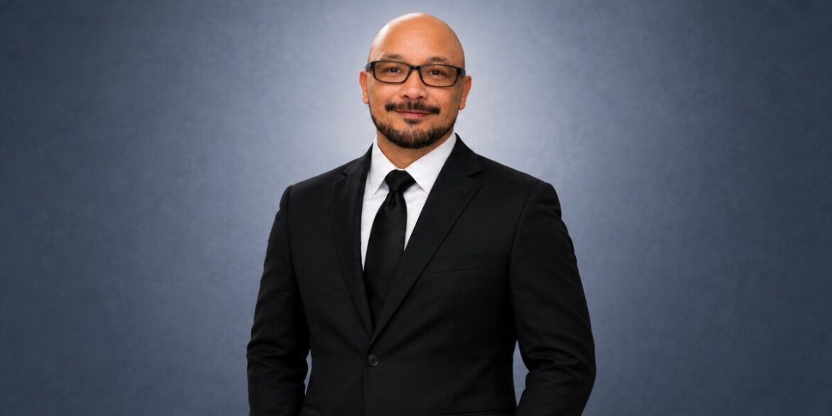 Professional portrait of Charles Santos wearing a black suit and tie against a gray background.
