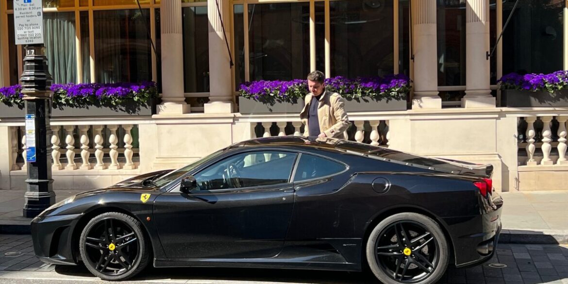 A man stands next to a black Ferrari sports car in London, showcasing elegance and professionalism in a city setting.