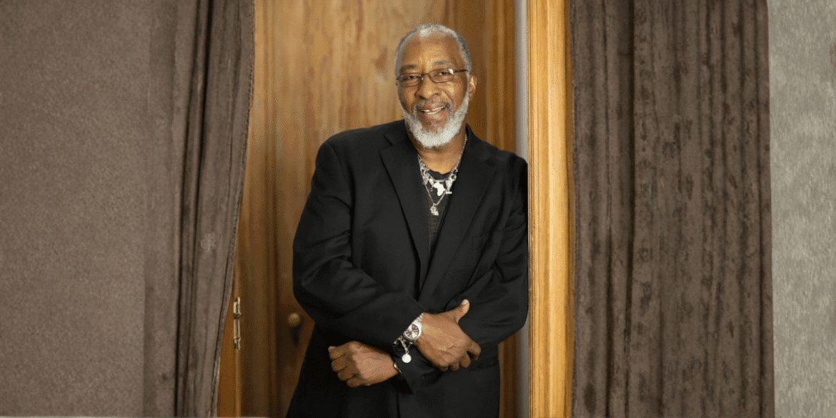 Smiling man with gray beard and glasses in black suit leaning in doorway between curtains.