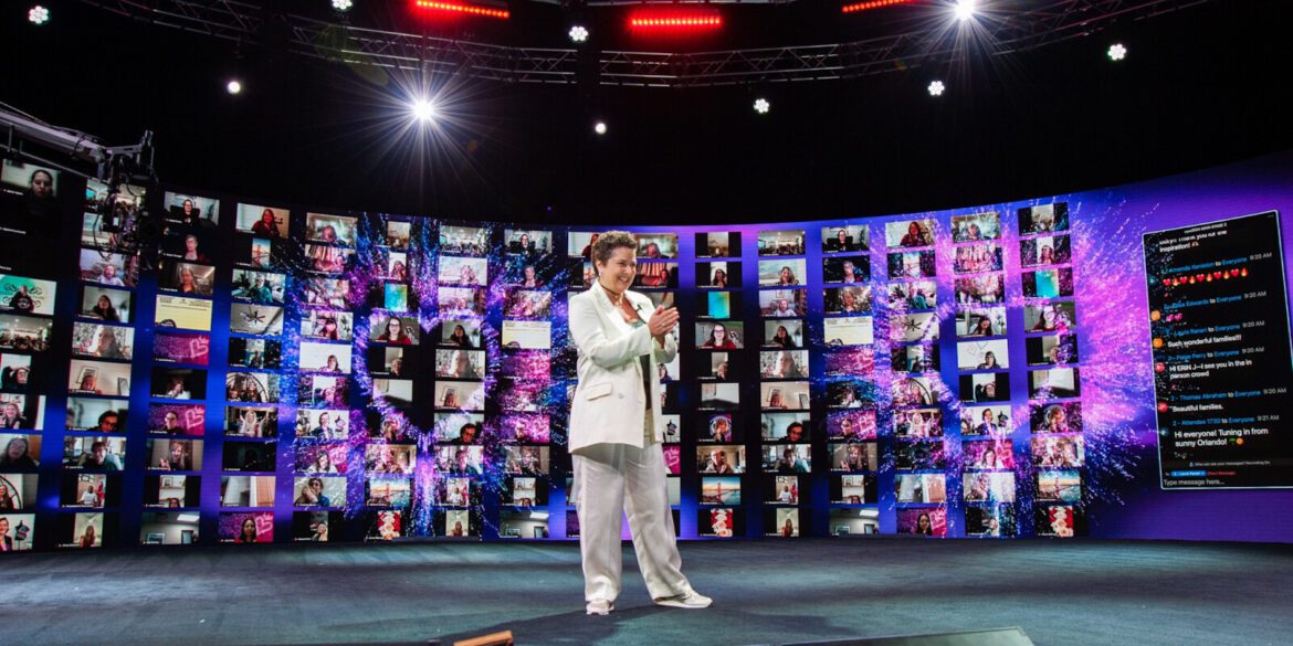 Josselyne Herman-Saccio speaking at an event, surrounded by virtual attendees displayed on large screens, celebrating her Evergreen Award.
