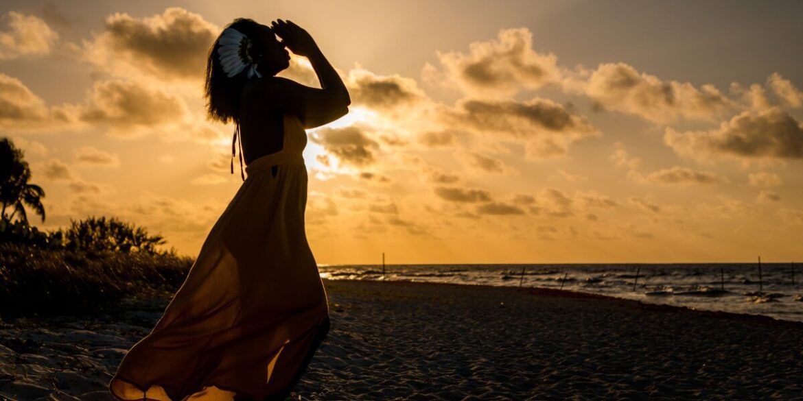 A woman with a flower in her hair stands on a beach at sunset, embodying peaceful reflection and global perspective.