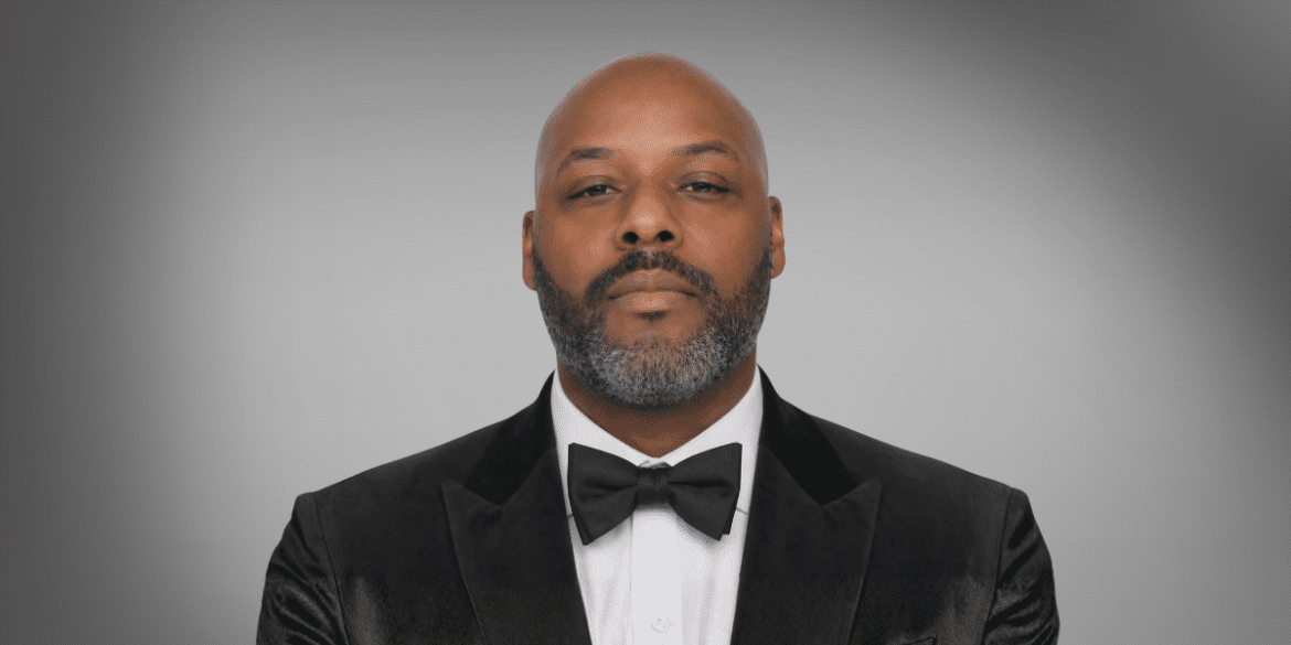 Man in black tuxedo and bow tie against gray background, formal portrait.