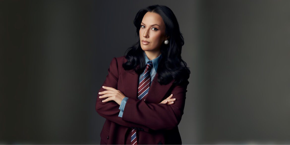 Confident woman in a tailored burgundy suit with a striped tie, exuding professionalism and leadership, arms crossed.