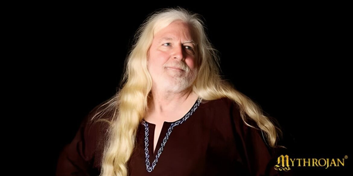A man with long white hair and a beard, Keith Wescovich, stands in a medieval workshop surrounded by crafting tools.