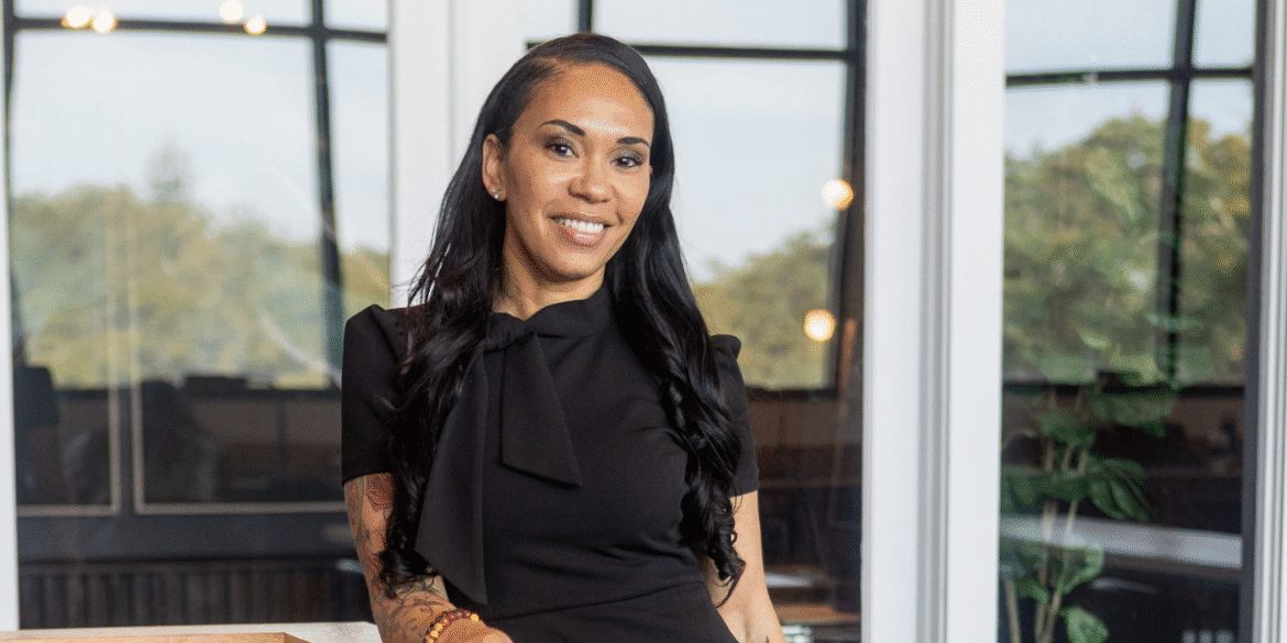 A woman in a black dress smiling confidently, standing in a bright, modern office with large windows and greenery in the background.