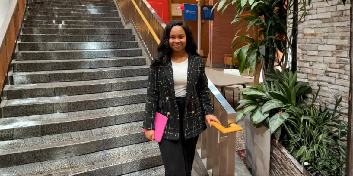 A professional woman in a checkered blazer and black pants stands confidently by a staircase, holding a pink tablet.