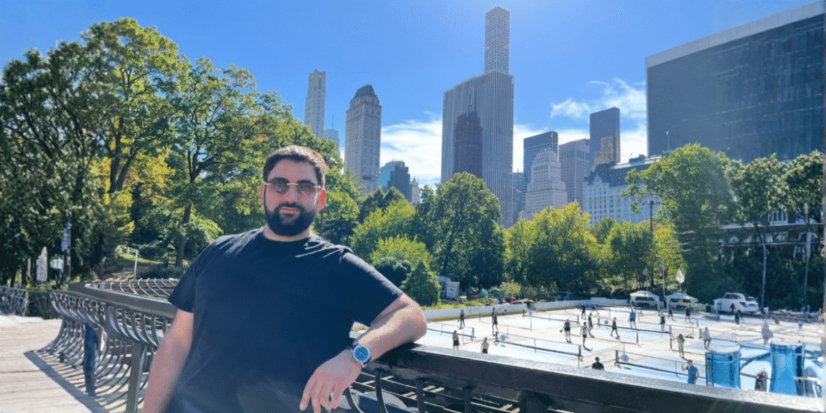 Alex Maghakian Gal leaning on railing in Central Park with Manhattan skyline behind.