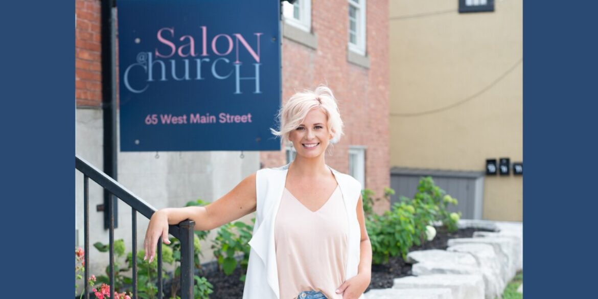 A smiling woman with short blonde hair stands outside a building with a sign reading "Salon@Church" on Main Street.