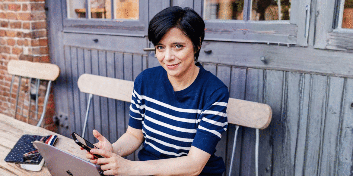 Woman using laptop and phone at outdoor café table, smiling confidently.