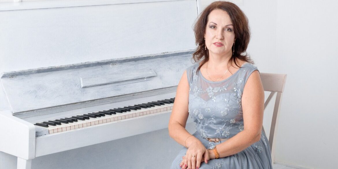 A woman in a blue dress sits on a chair beside a white upright piano, conveying a calm and composed atmosphere. The room is bright and minimalistic.