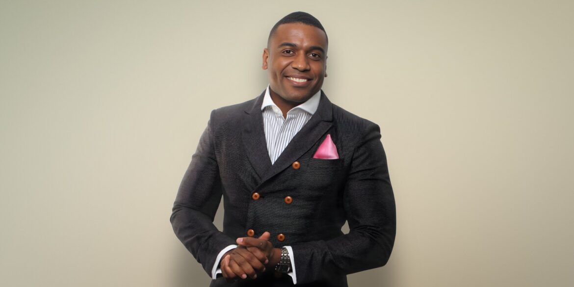 Man in a textured dark suit with a pink pocket square, smiling warmly against a light beige background. His hands are clasped in front.