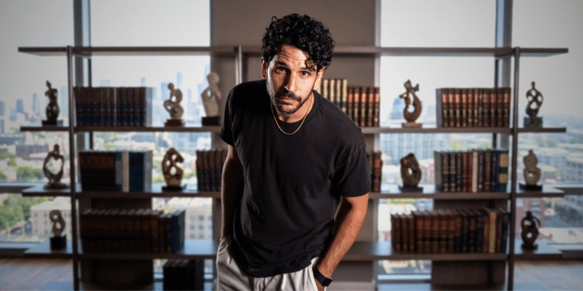 Man with curly hair, wearing a black t-shirt, standing in front of a bookshelf with a city view.