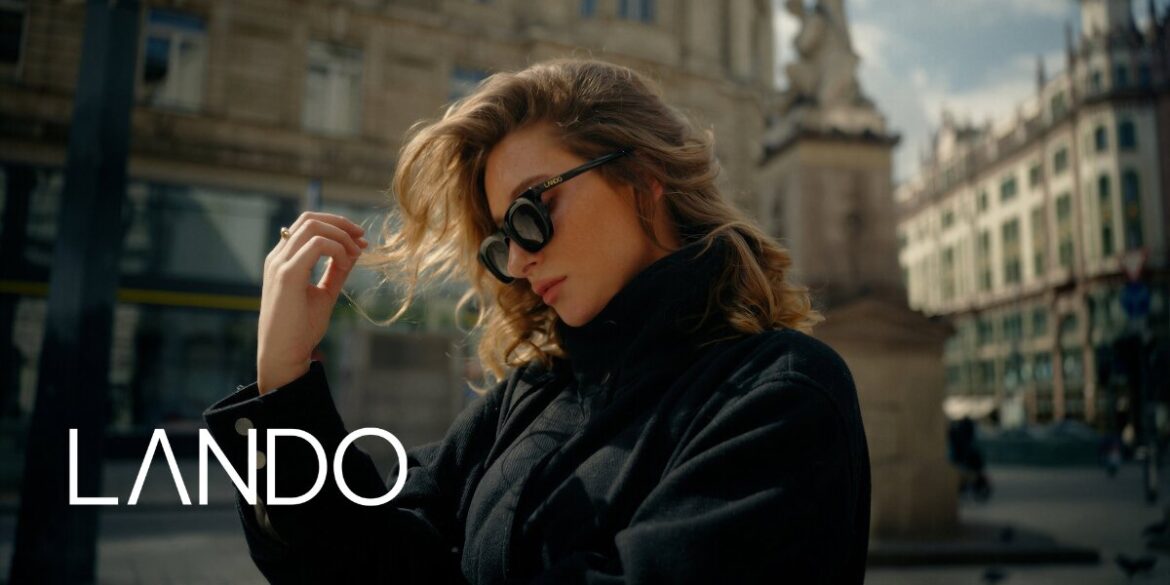 A woman in sunglasses and a black coat stands in a city square, with elegant architecture behind her. She looks contemplative. The word "Lando" is displayed.