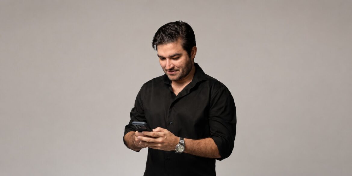 Man in black shirt looking at smartphone, wearing a wristwatch, against a neutral studio background.