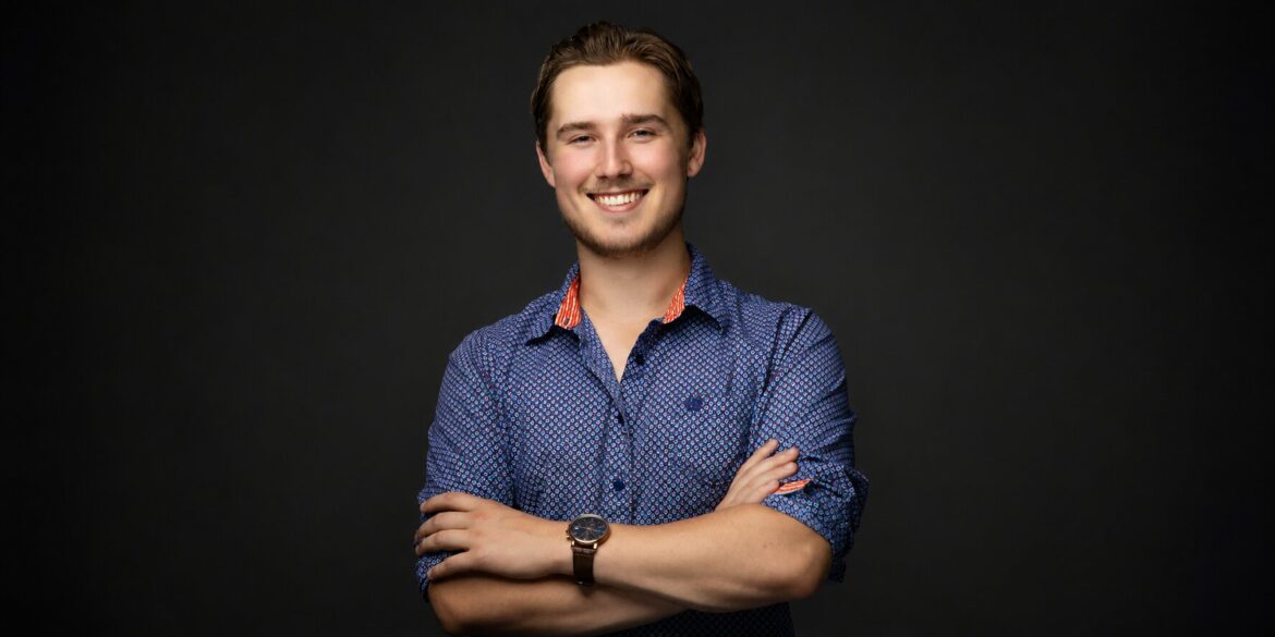 Professional portrait of Trystan Keller smiling with arms crossed against a dark background.