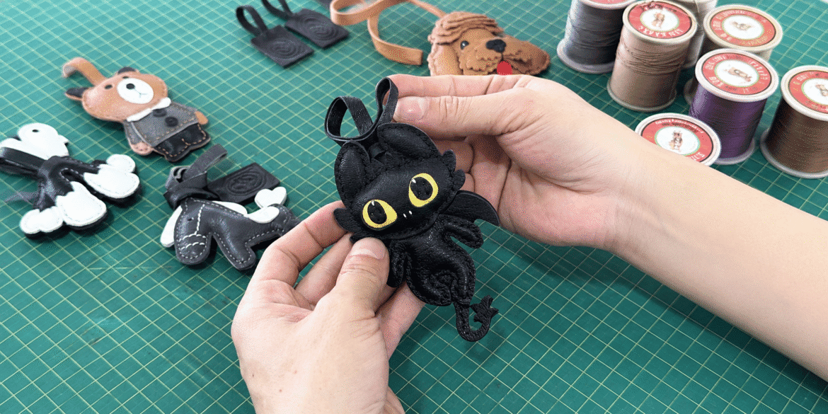 Hands holding a handcrafted black leather dragon charm on a workshop cutting mat.