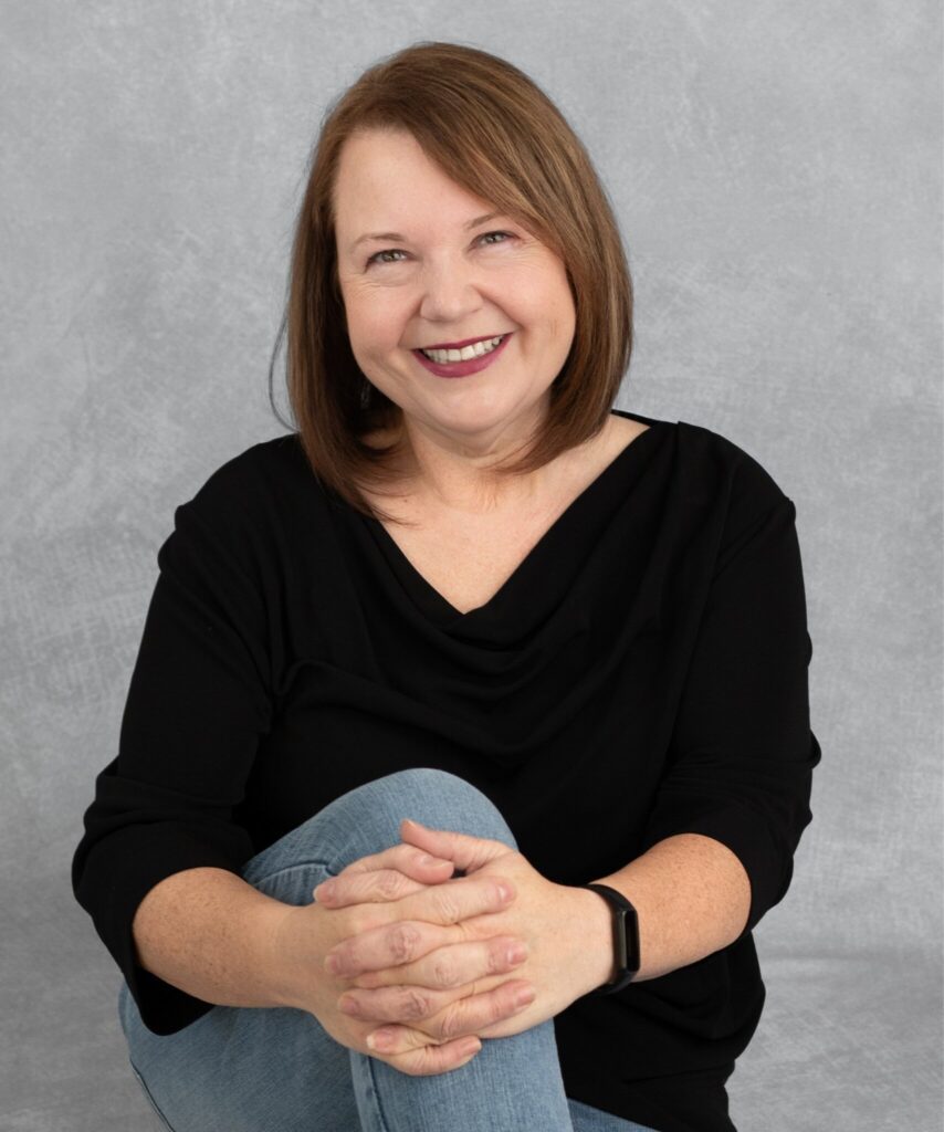 Christina Bedal smiling while seated casually against a gray backdrop, wearing a black top and jeans.