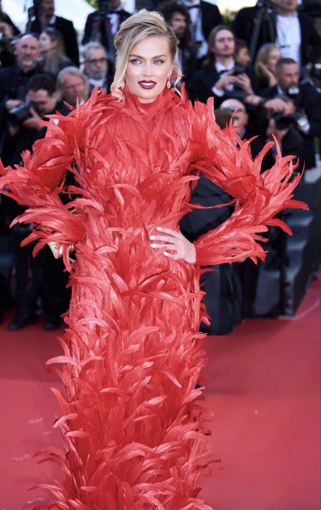 Woman wearing a dramatic red feathered gown posing on a red carpet before photographers.