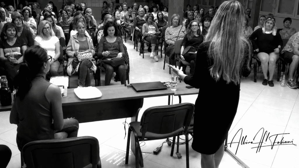 Speaker addresses a large audience during a healthcare education seminar, with attendees listening attentively in a conference room.