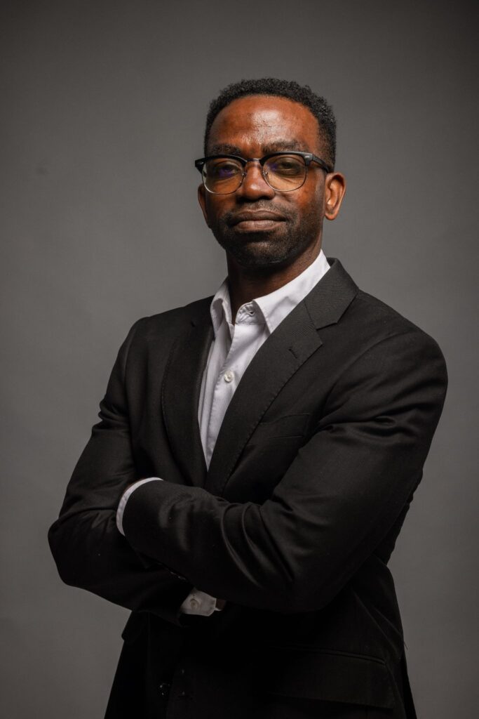 Christopher Haywood in a black suit with folded arms against a gray studio background.