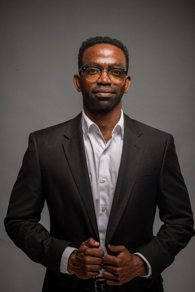 Christopher Haywood wearing a black suit and white shirt in a studio portrait.