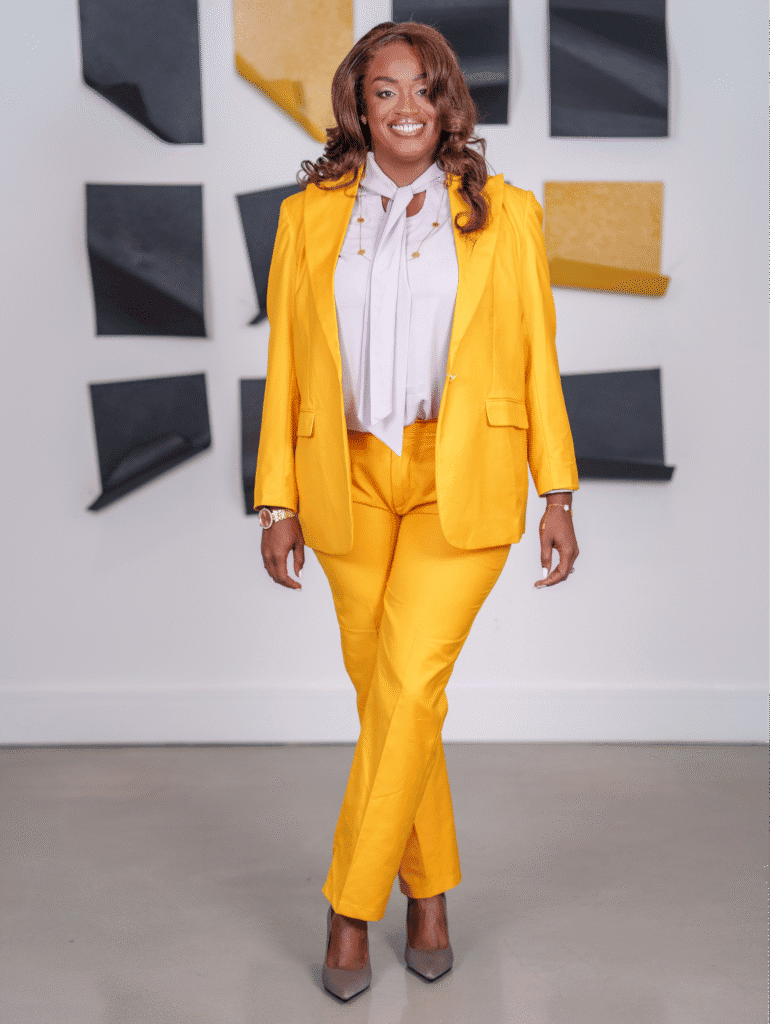 Woman in bright yellow suit smiling confidently in front of abstract black and gold wall art.