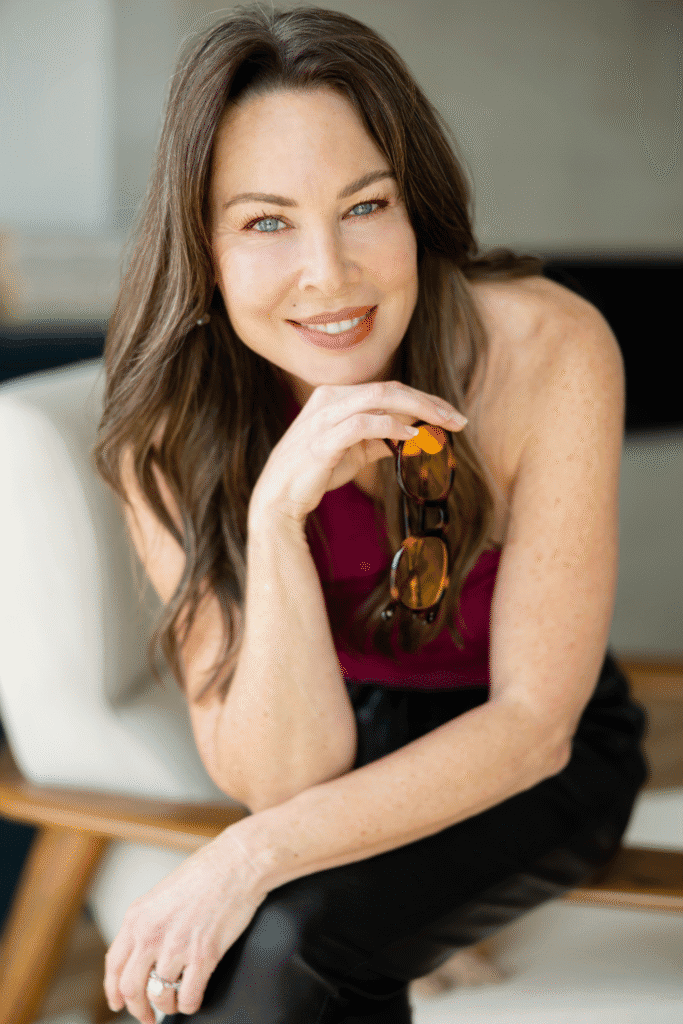 Kim Wheeler smiling while holding sunglasses during an indoor portrait.