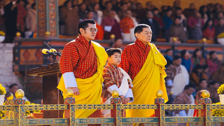 Three Bhutanese individuals in traditional attire stand at a ceremonial podium.